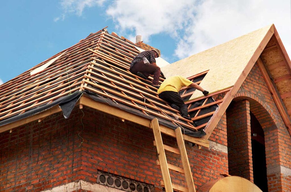 Two workers carefully installing a tarp and plywood sheets to shield a sloped residential roof during renovation.