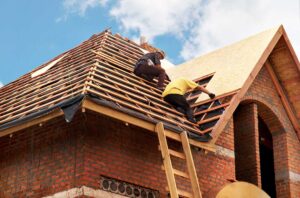 Two workers carefully installing a tarp and plywood sheets to shield a sloped residential roof during renovation.
